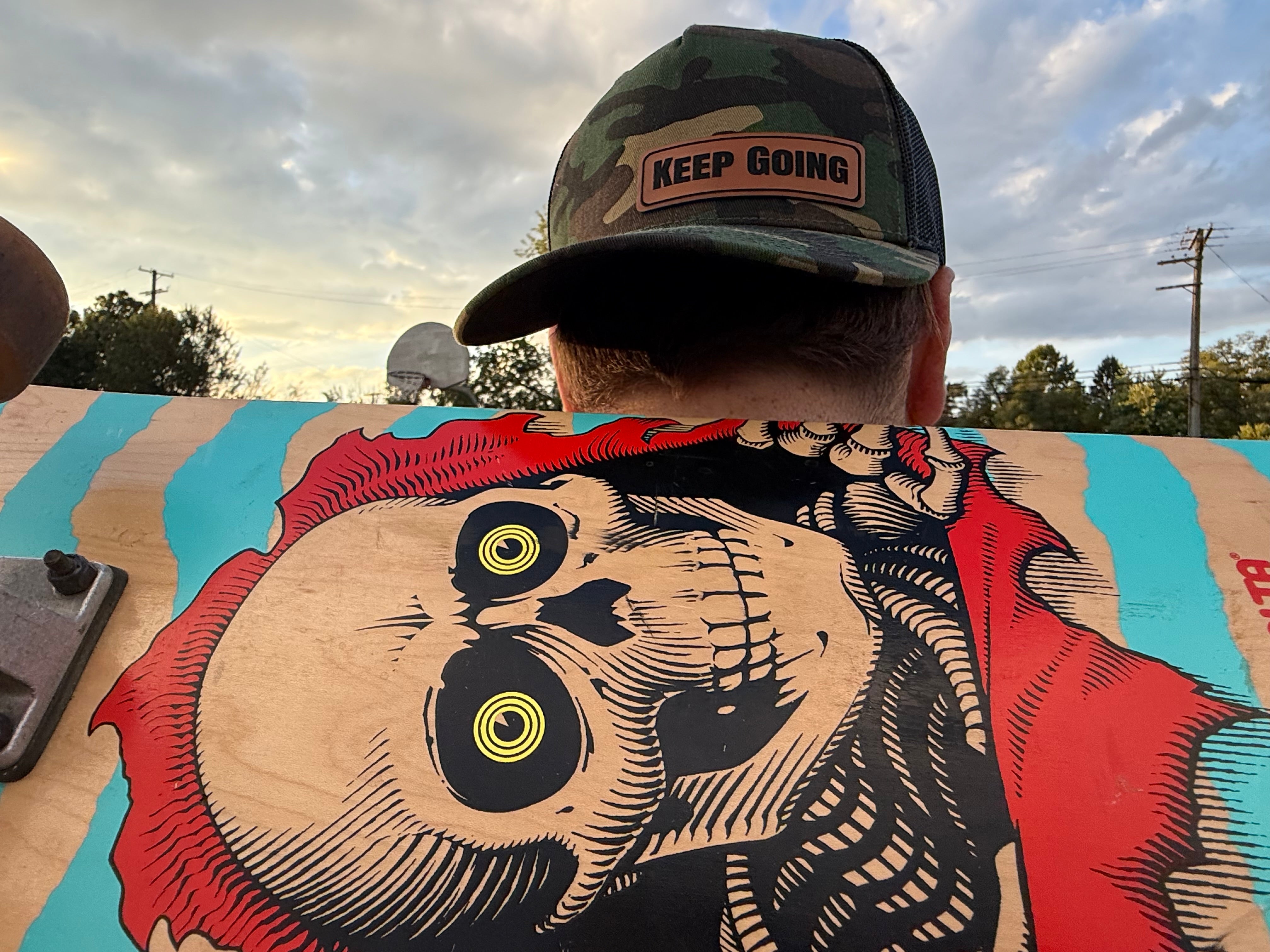 Person wearing a cap with 'Keep Going' patch, sitting on a skateboard with a skull and owl design.