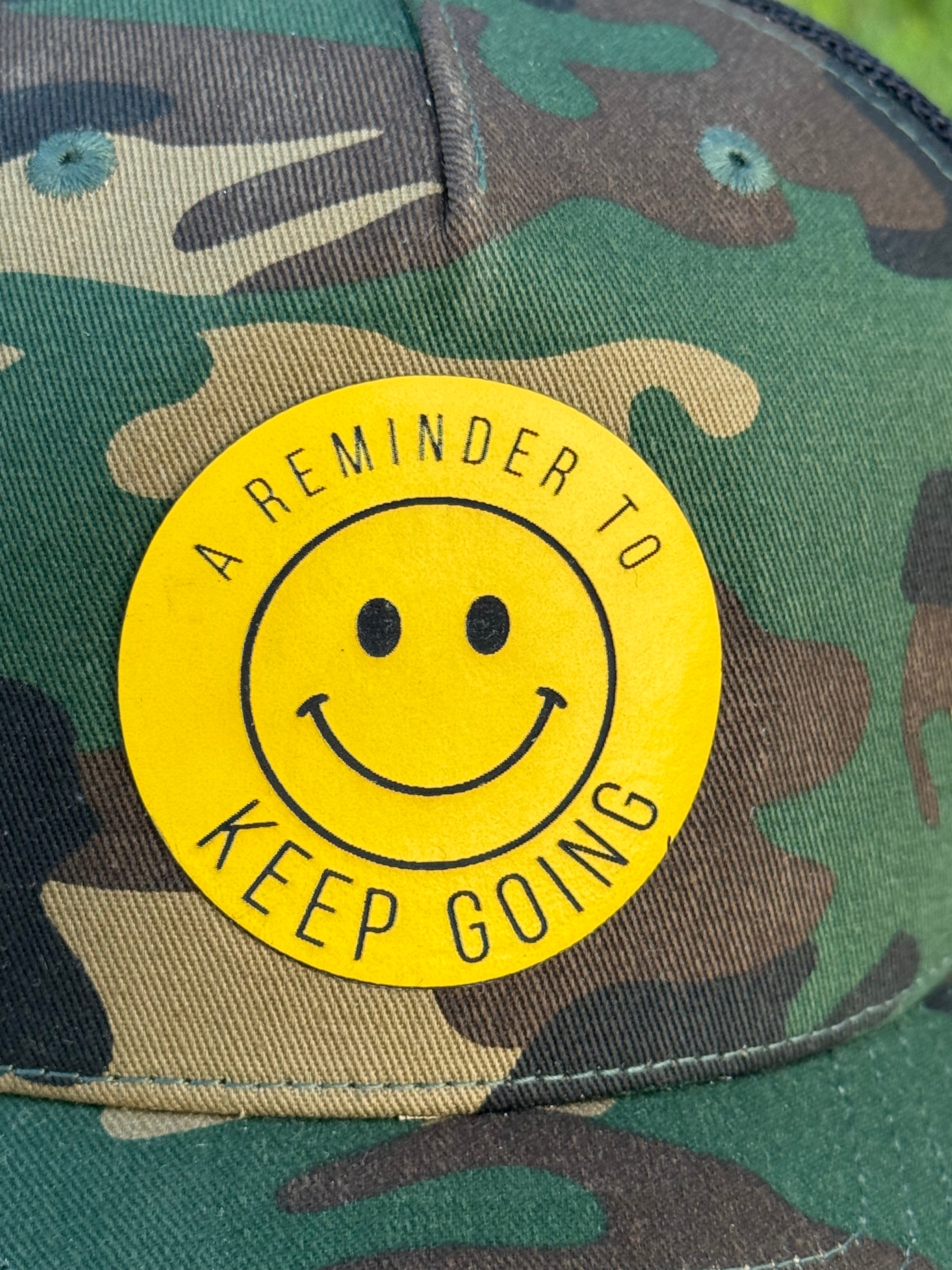Camouflage cap with a yellow circular patch featuring a smiley face and text.