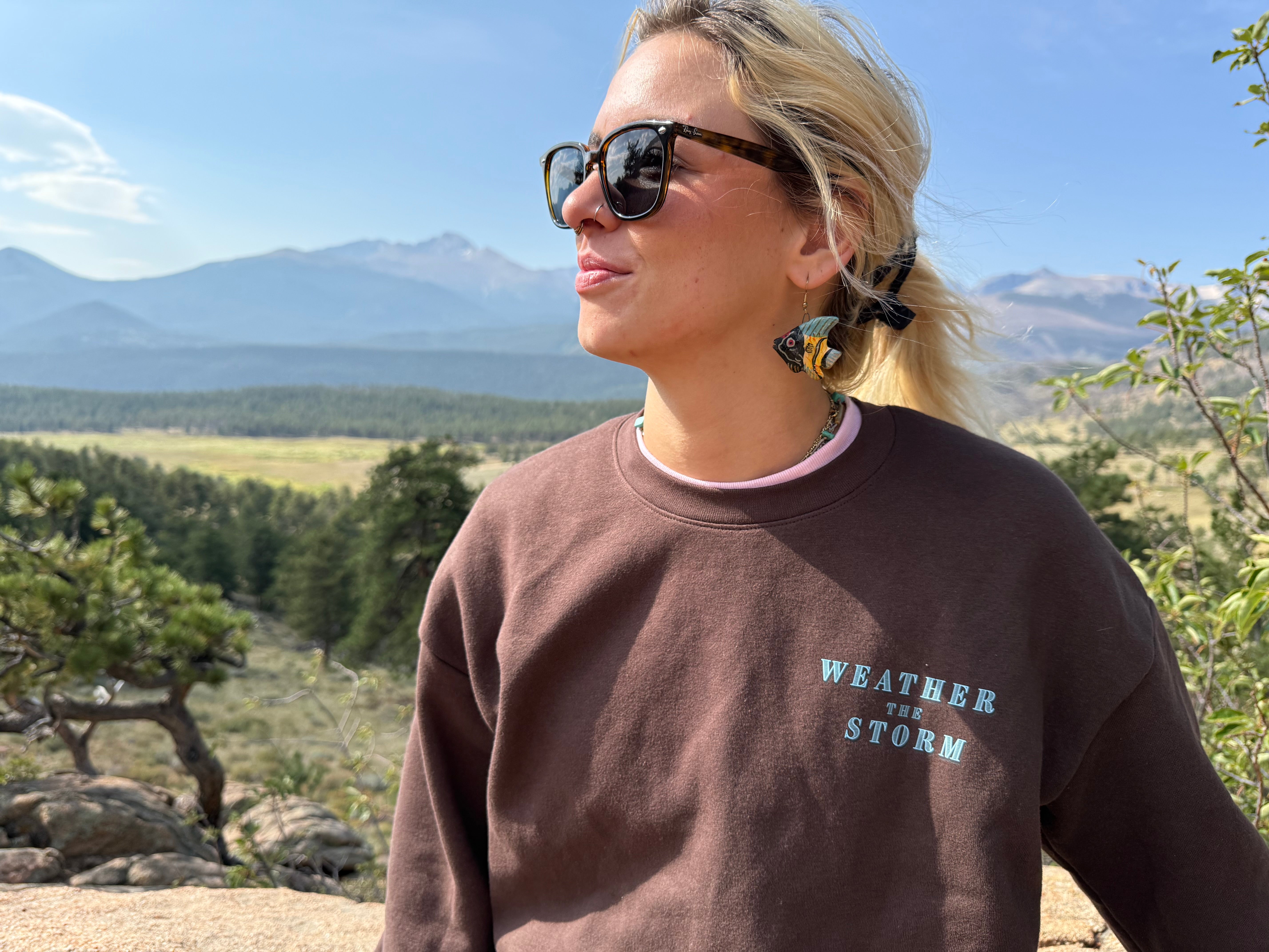 Person wearing sunglasses and a 'Weather the Storm' sweatshirt in a natural setting with mountains and trees.