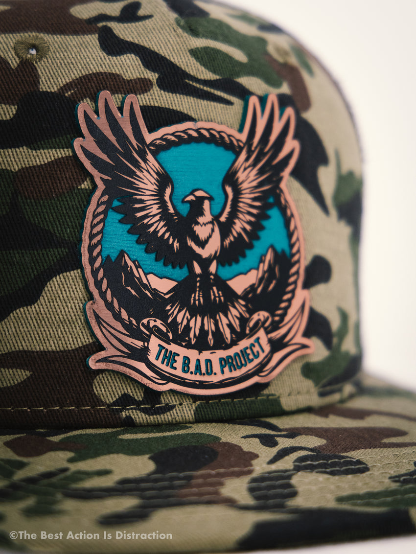 Camouflage cap with an eagle emblem and 'The B.A.D. Project' text on a blurred background.