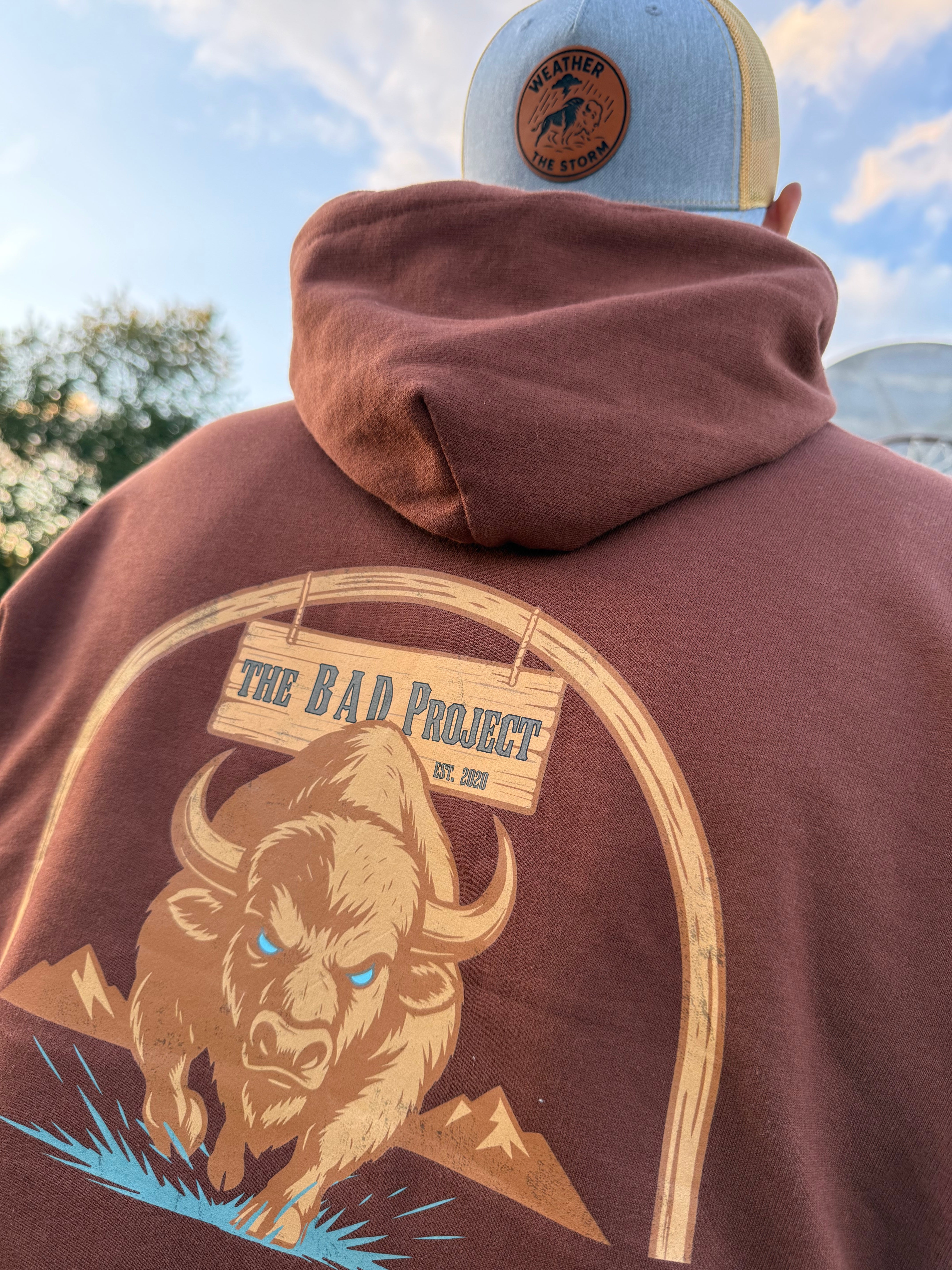 Brown hoodie with bear graphic and 'The Bad Hoods' text, worn by a person outdoors.
